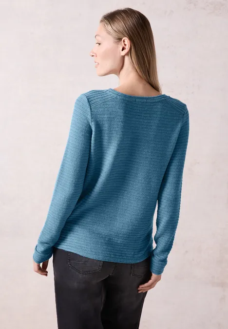 Frau mit blauem Pullover und dunkler Hose steht seitlich und schaut nach links.